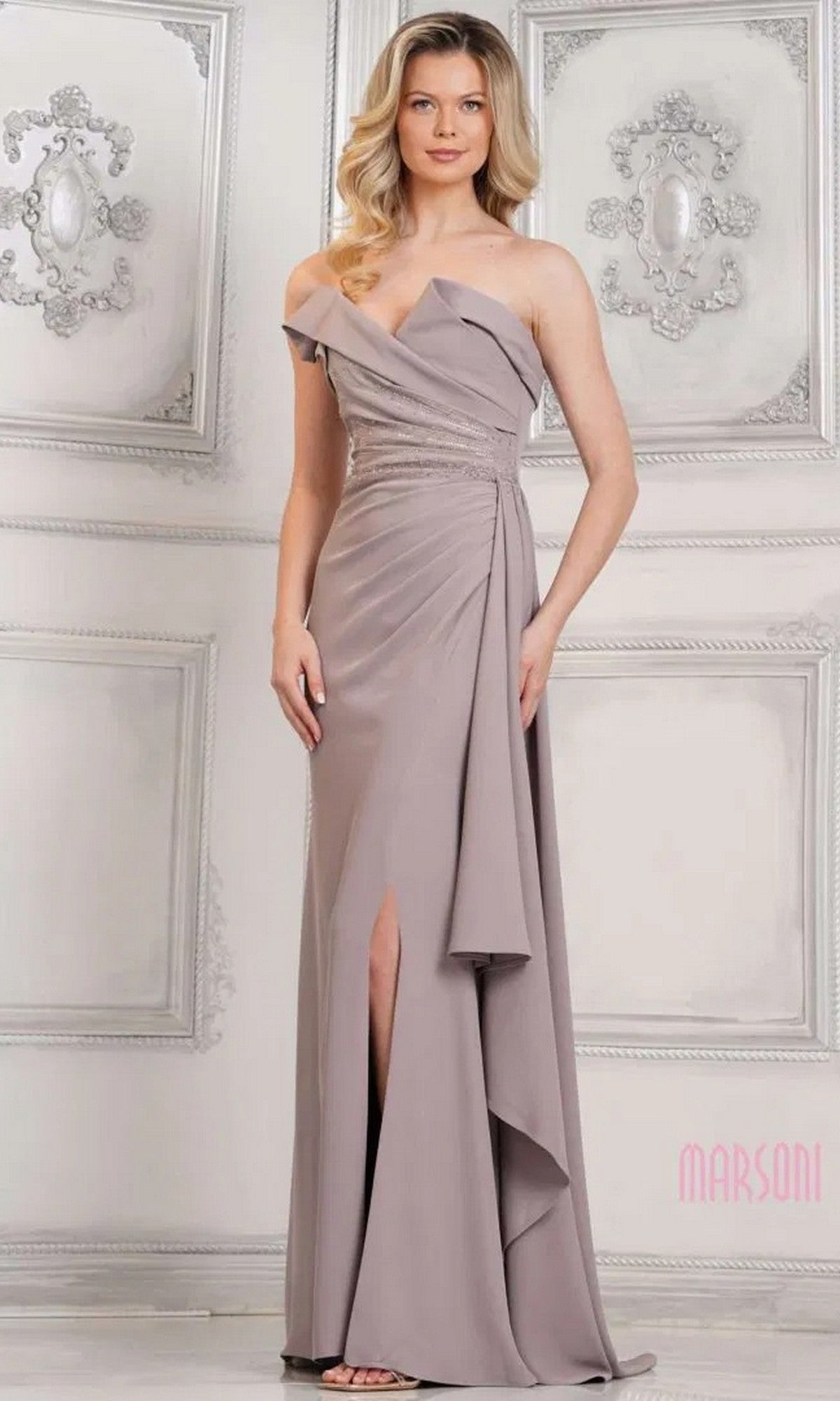 A woman stands indoors wearing the Marsoni MV1304, a taupe, floor-length formal dress with a beaded bodice, side slit, draped detailing, and off-the-shoulder design. Ornate white wall panels form the background.